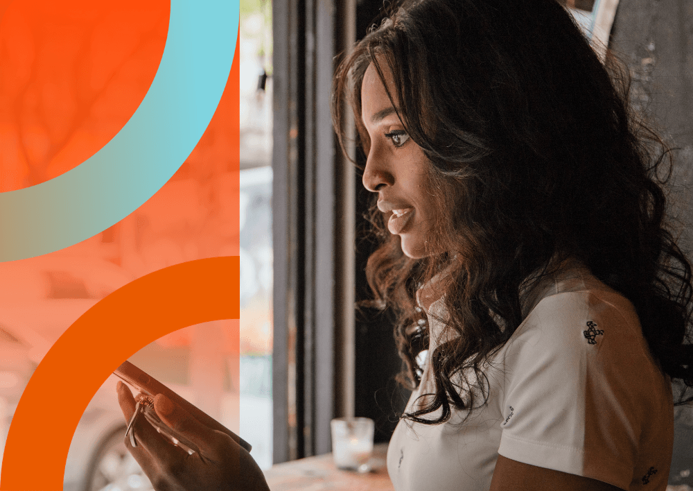 Smiling woman with glasses and backpack holds a notebook, sitting on steps. Another person reads in the background.